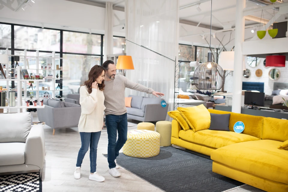 Un couple dans un magasin de mobilier moderne examine des canapés, l'homme désignant un modèle jaune tandis que la femme l'écoute attentivement