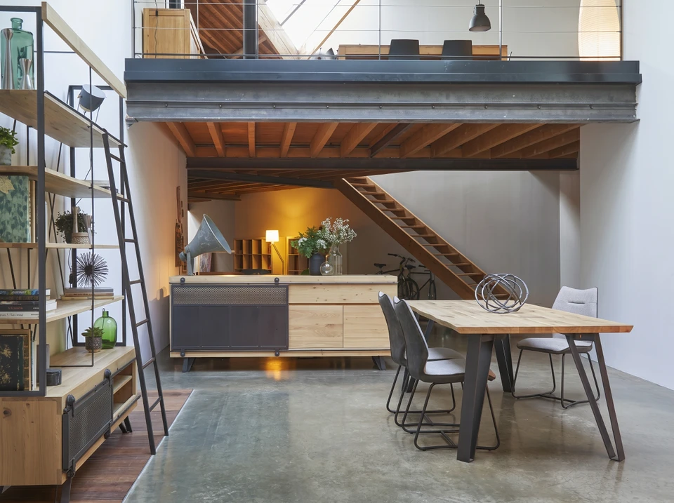 Personne tenant une tablette tactile affichant un canapé gris avec coussins orange en réalité augmentée dans un intérieur domestique minimaliste aux murs blancs et parquet bois clair